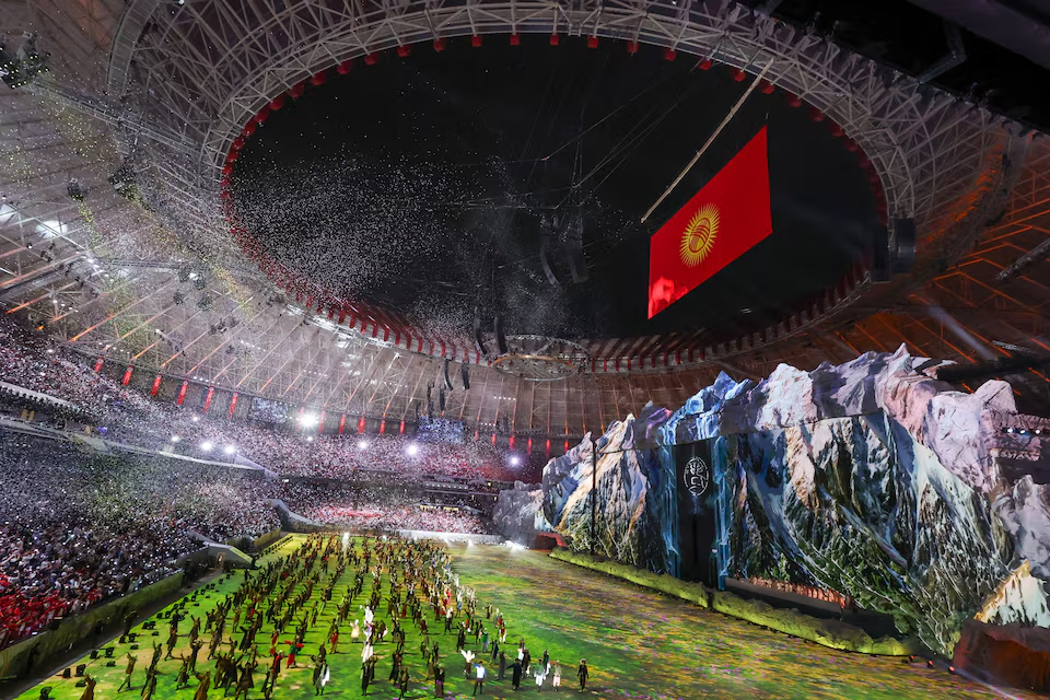 Performers fill a packed stadium field during a World Nomad Games ceremony with a large Kyrgyzstan flag hanging over a mountain stage
