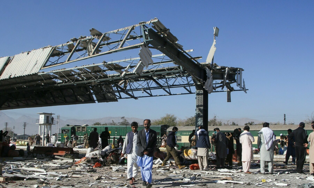 People walking through scattered debris at a train station platform under a severely damaged metal roof structure