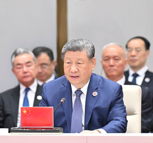 Xi Jinping speaks into a microphone at a conference table with a small Chinese flag in front of him
