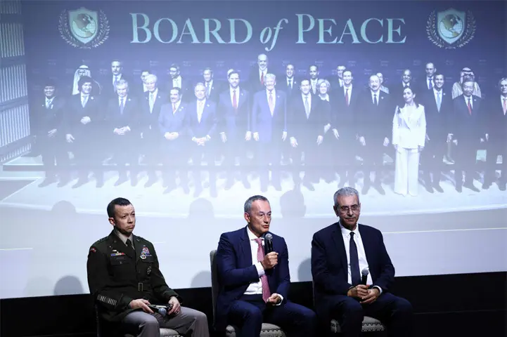 Three speakers, including a military officer, sitting on stage in front of a screen reading Board of Peace