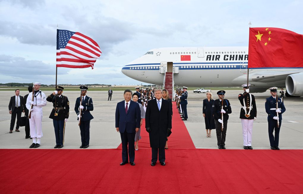Xi Jinping and Donald Trump stand on a red carpet at an airport, flanked by honor guards holding US and Chinese flags