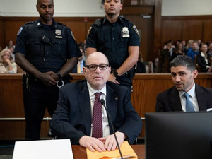 Harvey Weinstein sits at a defense table in a courtroom beside his attorney, with two police officers standing behind him