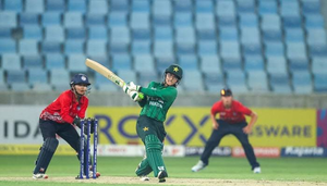 A Pakistani women's cricket player in a green uniform swings her bat during a match near the wicketkeeper