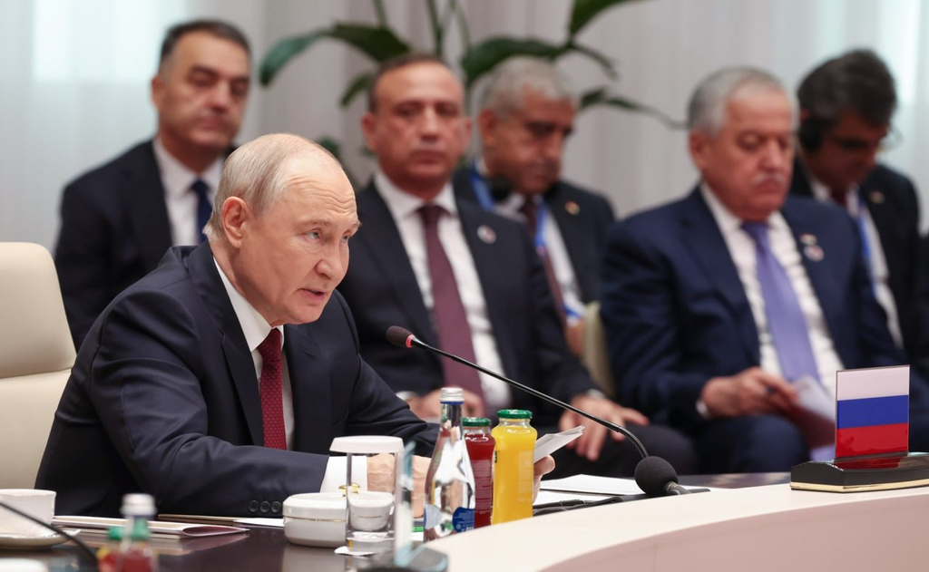 Vladimir Putin speaks into a microphone at a conference table with delegates seated behind him